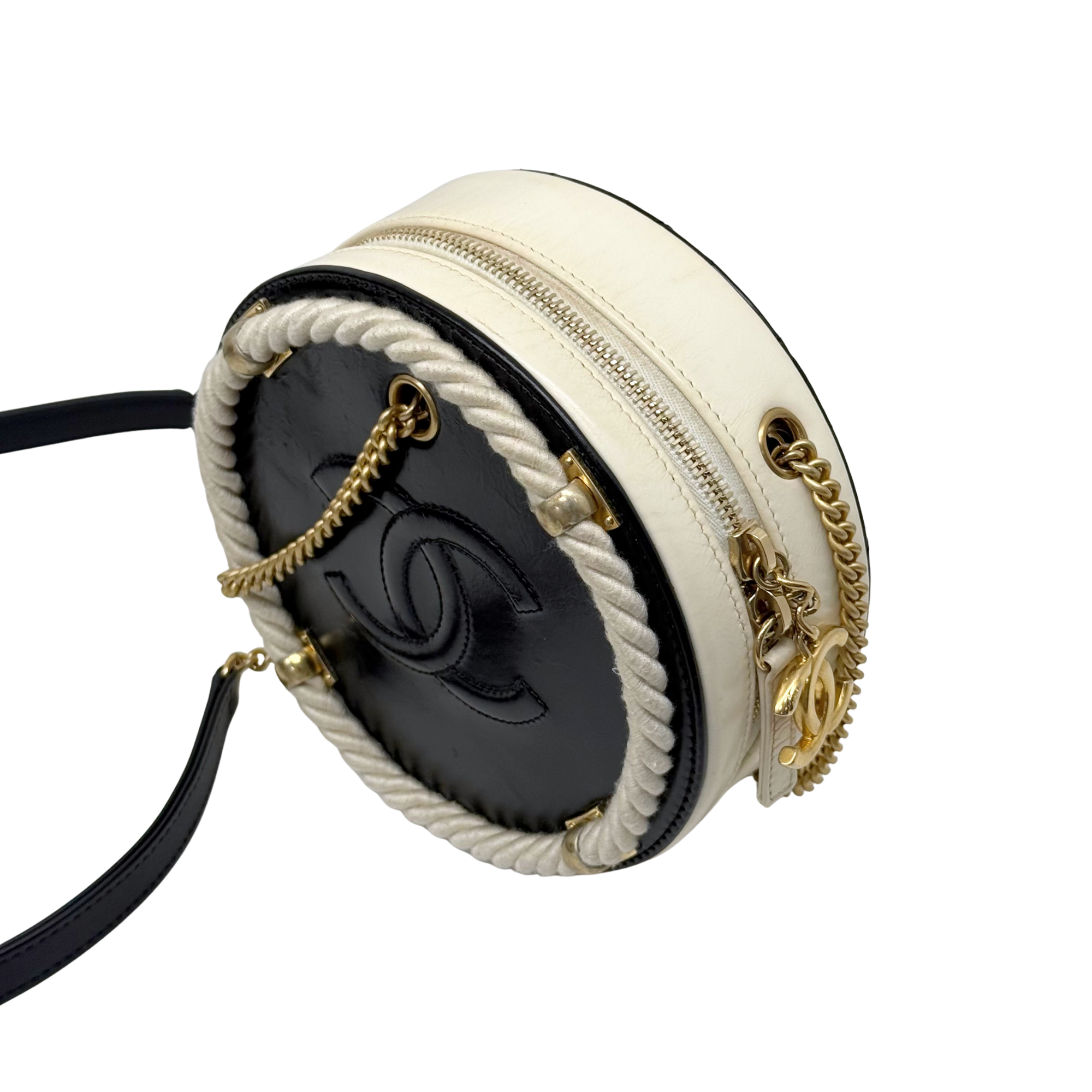 Round cream mini crossbody bag with black interior, braided trim, and gold chain strap.